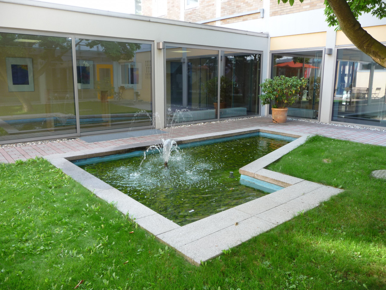 Bild eines Innenhofes der Hoffmanns Höfe zu sehen ist ein Teich mit Wasserspiel und eine grüne Wiese sowie die Fensterfront.